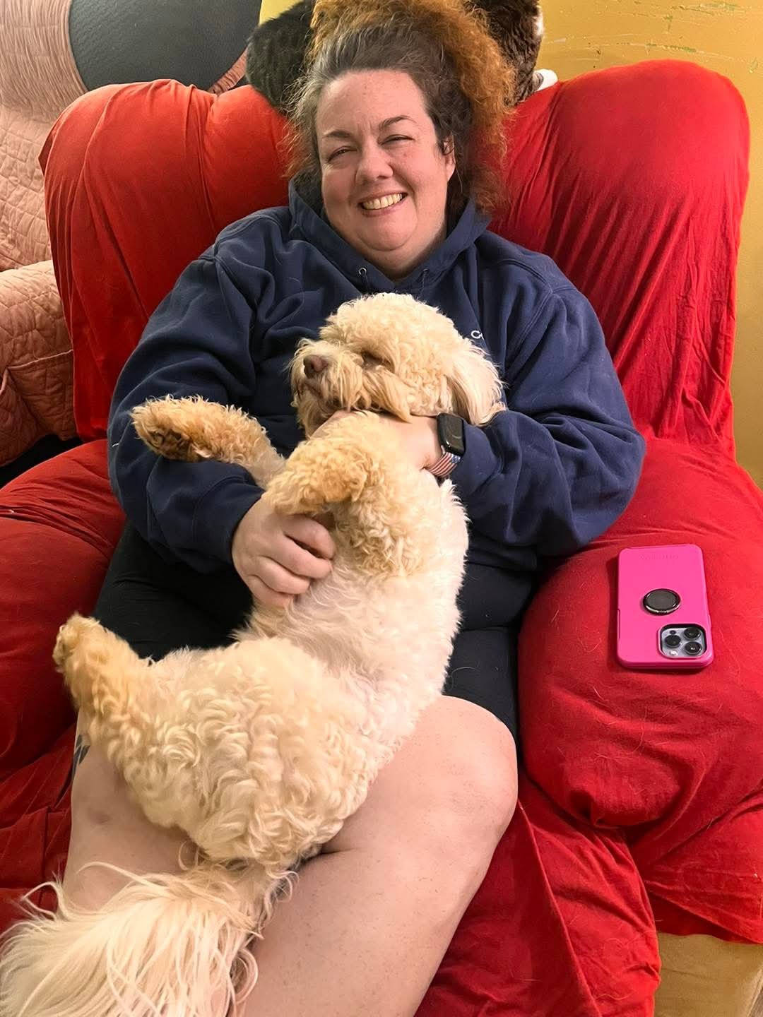 Kerri, Owner & Founder of Doods & Dolls, holding a fluffy beige dog on a red couch